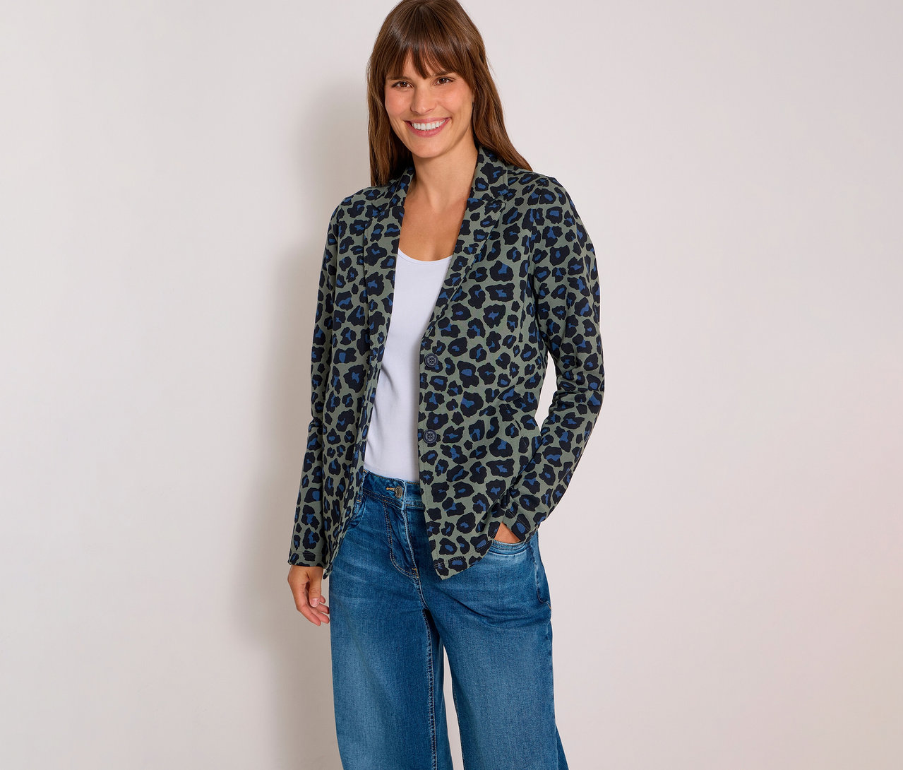 Lächelnde Frau in einem grünen CECIL Jerseyblazer mit Leopardenmuster, weißem Top und blauen Jeans, Hände in den Taschen.