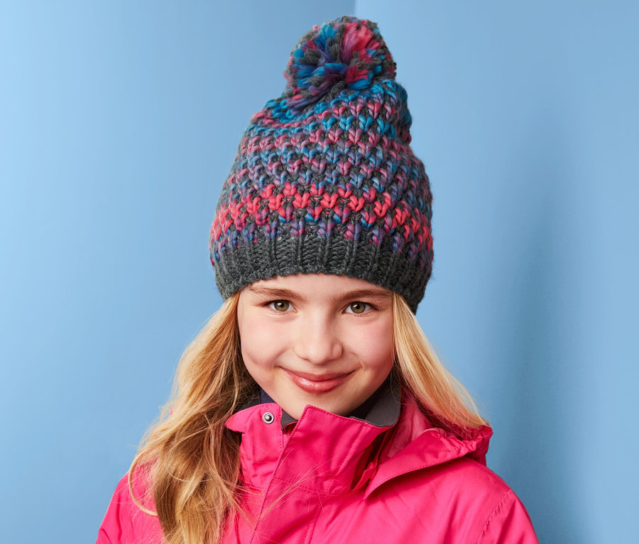 Mädchen mit blonden Haaren und Strickmütze trägt eine pinke Jacke vor blauem Hintergrund.