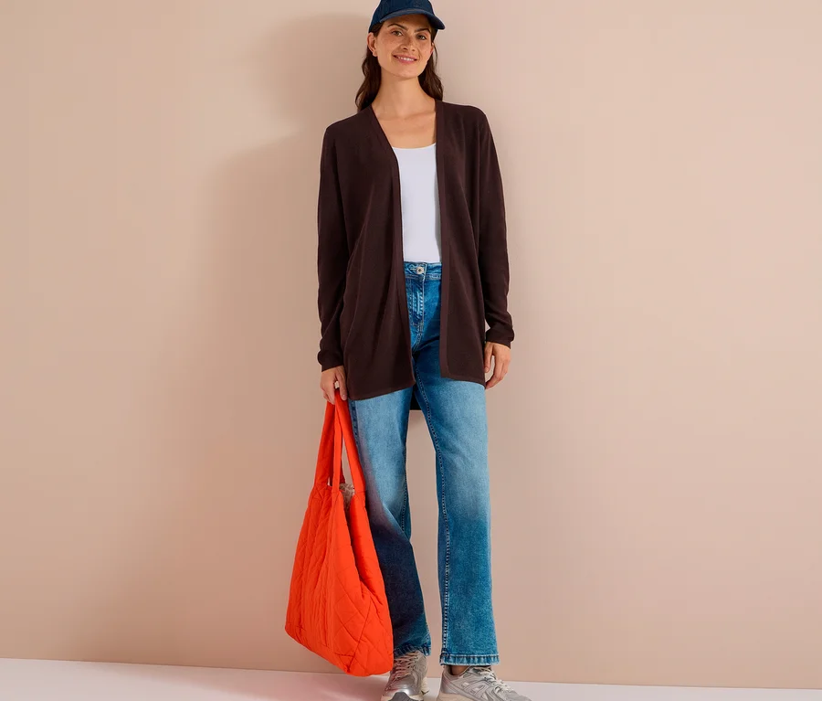 Frau steht lächelnd, trägt eine dunkelbraune CECIL Strickjacke, weißes Top, Jeans, blaue Kappe und hält eine orangefarbene Stepptasche.