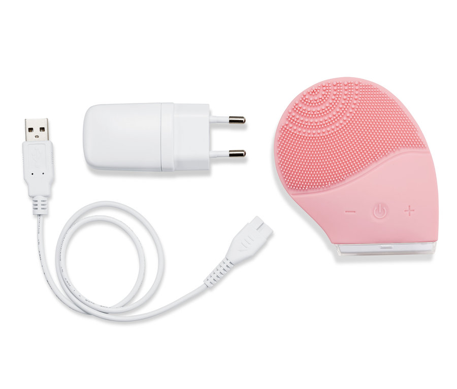 Eine elektrische Silikon-Gesichtsreinigungsbürste in Rosa liegt mit Ladekabel und Adapter auf weißem Grund.