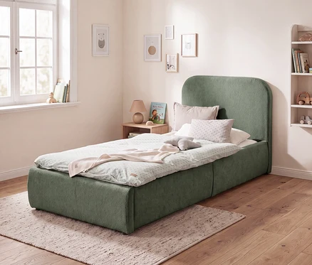 Ein Kinderzimmer mit einem grünen Polsterbett mit abgerundetem Kopfteil, bedeckt mit einer hellgrünen Bettdecke, einer rosa Decke und Kissen. Daneben steht ein Nachttisch mit Lampe und Buch. An der Wand hängen Bilder, und ein Regal mit Spielzeug ist sichtbar.