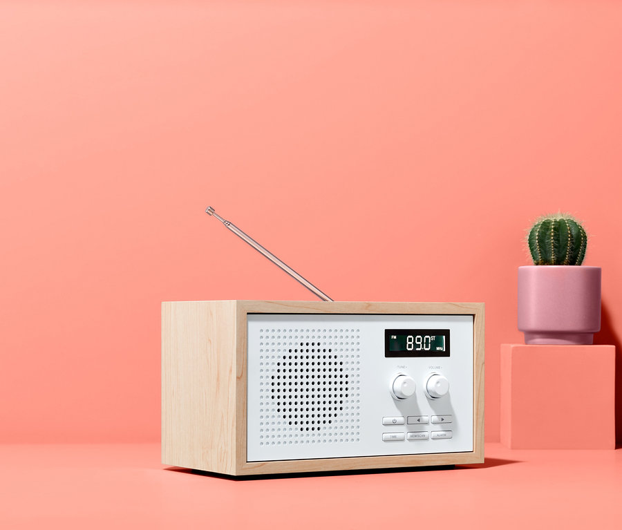 Design-FM-Radio im formschönen Holzgehäuse neben kleinem Kaktus in rosa Topf auf korallenrotem Hintergrund.