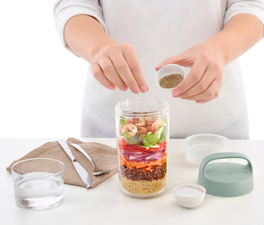 Eine Frau würzt eine To-go-Lunchbox, die mit Salat-Schichten gefüllt ist.