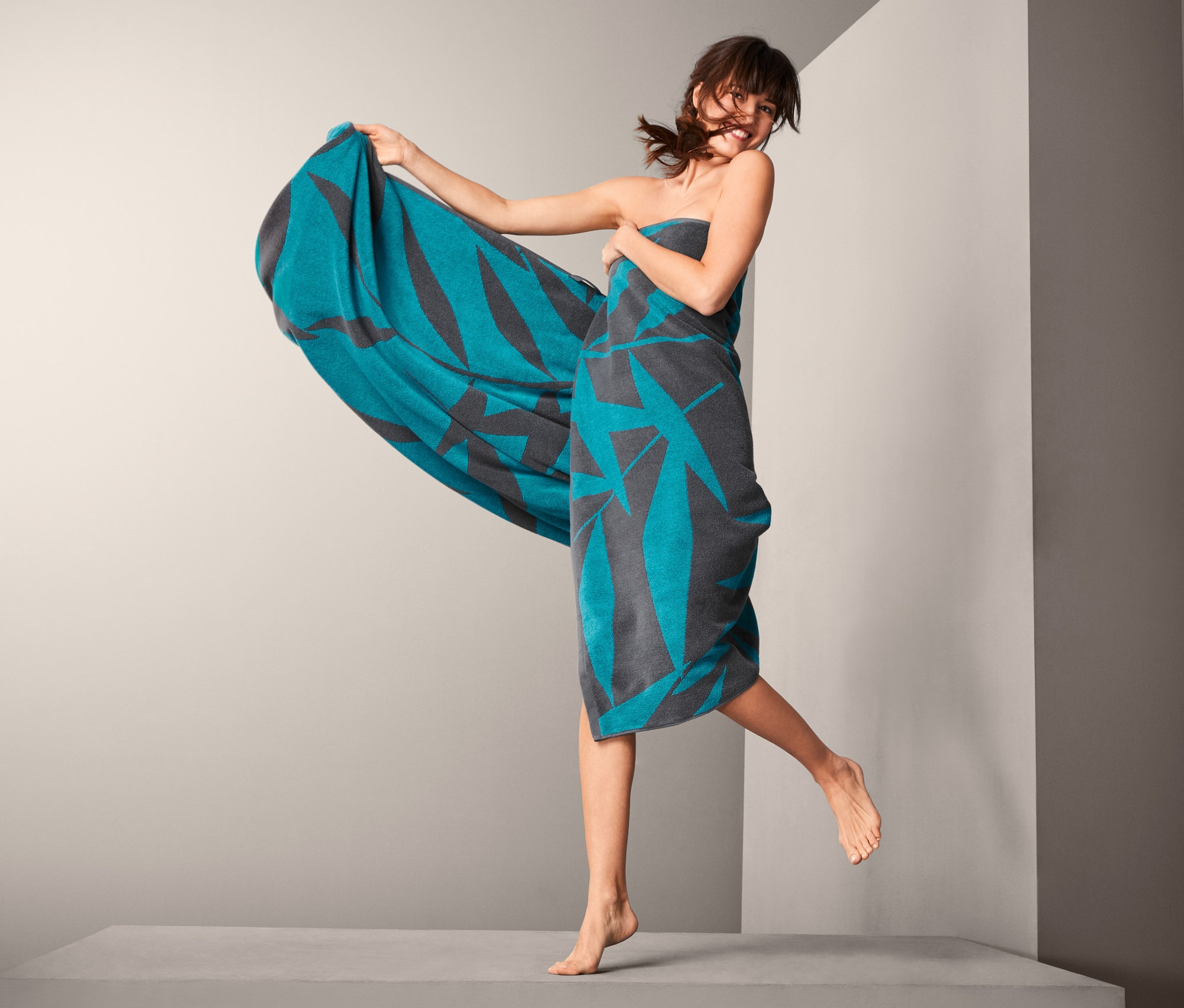 Frau springt, eingewickelt in ein grau-türkises XL-Frottier-Badetuch.
