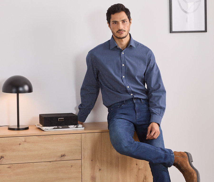 Mann sitzt auf einer Holzkommode und trägt ein blaues Hemd, Comfort Fit.
