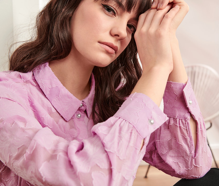 Frau mit braunen Haaren hat die Hände am Gesicht und trägt eine helllila Hemdbluse mit Strukturmuster.