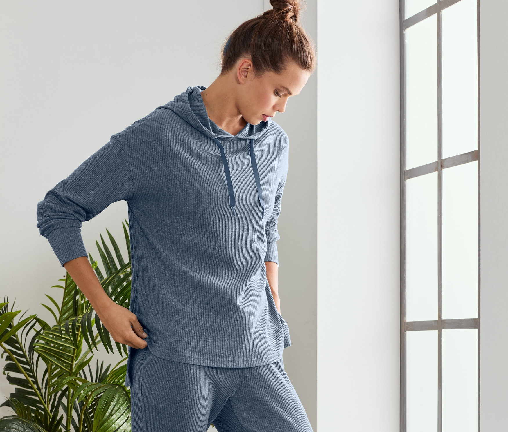 Frau im blauen Relaxshirt steht an einem Fenster.