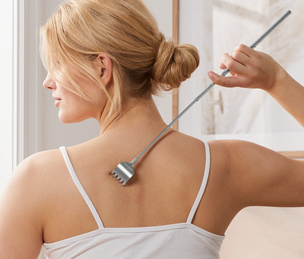 Frau sitzt im Bett und kratzt sich mit einem Rückenkratzer am Rücken.