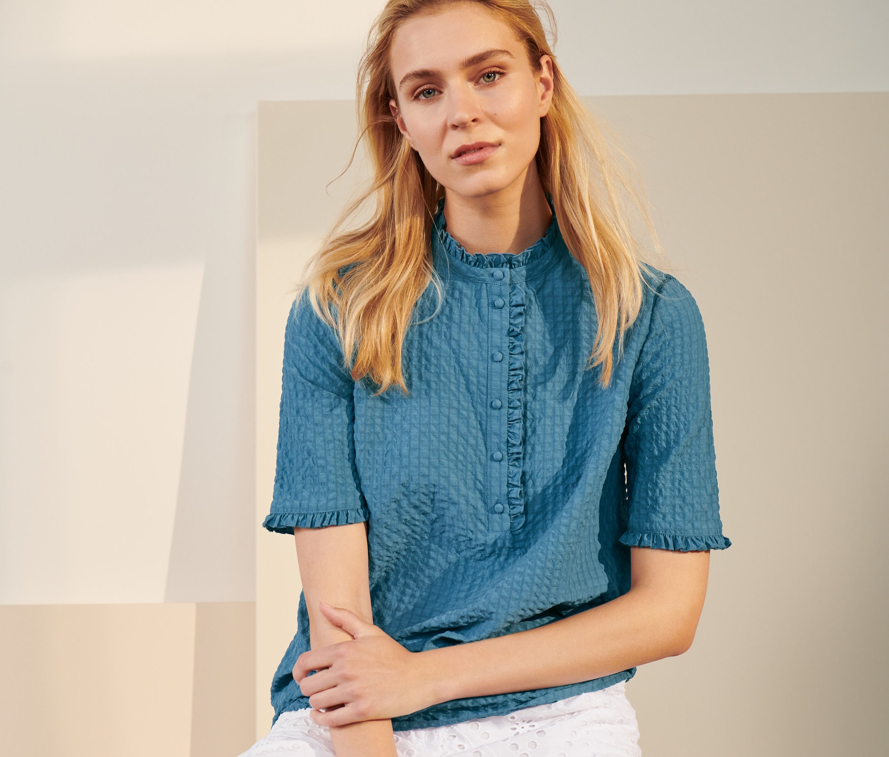 Frau sitzt auf gelbem Hocker und trägt eine blaue Bluse in Crinkle-Optik und weiße Shorts.