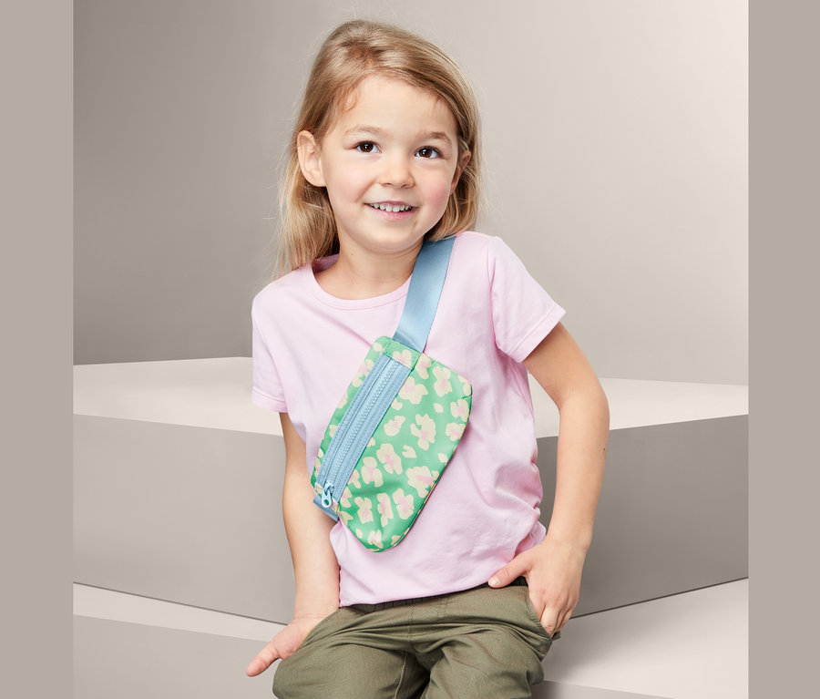 Lächelndes Mädchen sitzt und trägt eine grüne Bauchtasche mit Print.