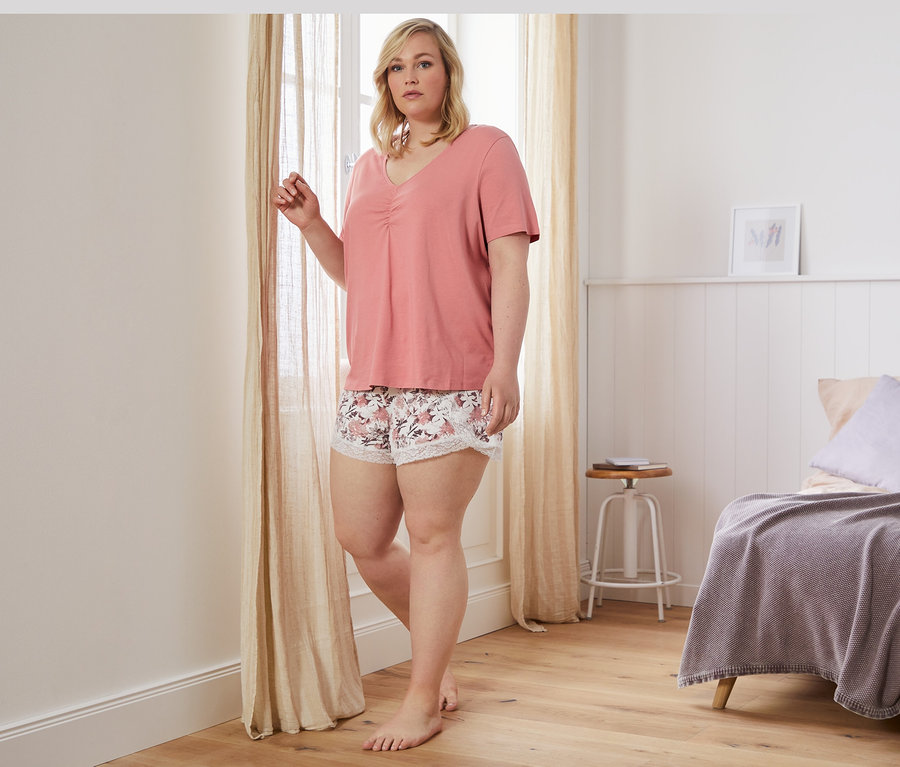 Frau steht am Fenster und trägt einen Shorty-Pyjama.