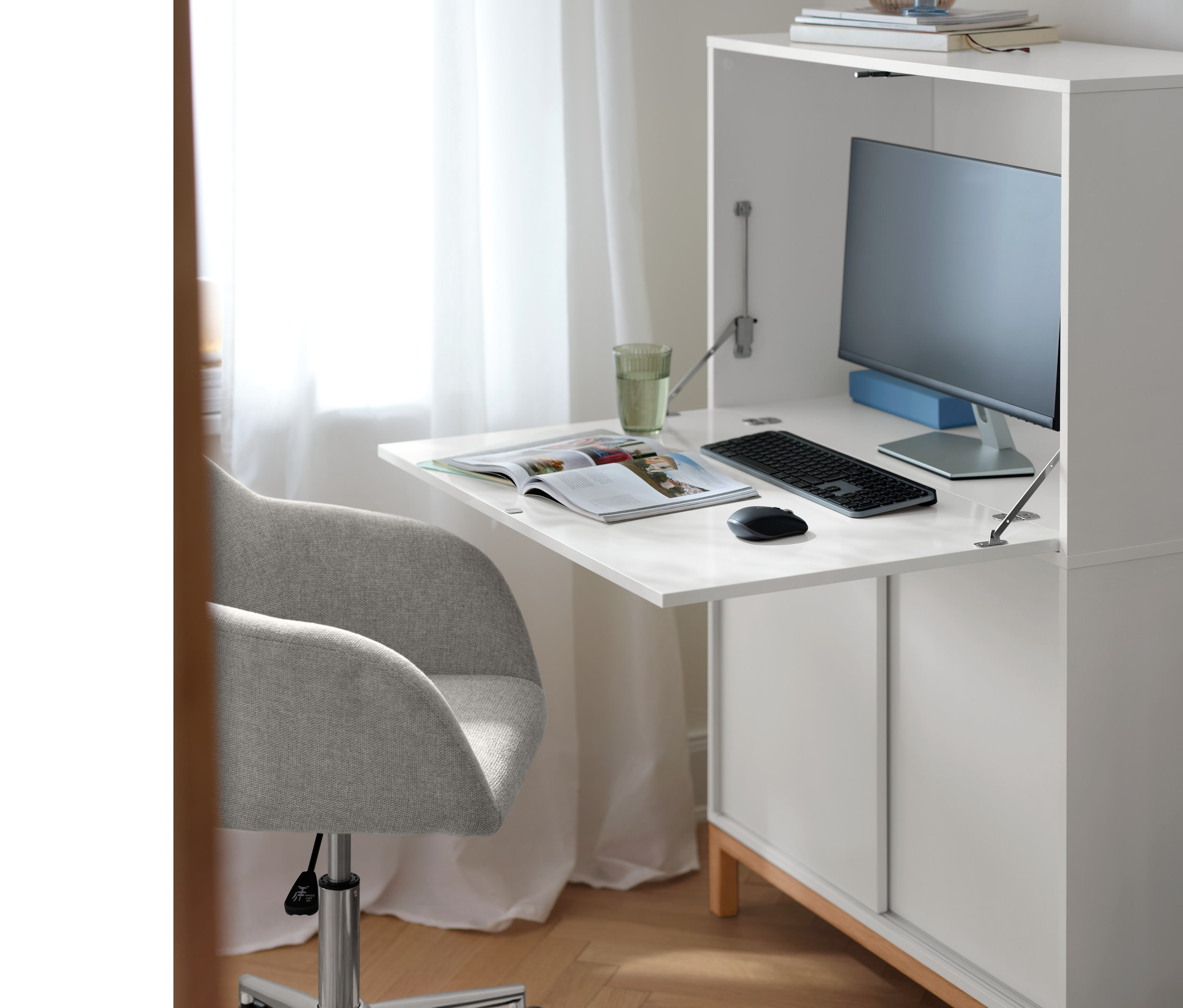 Beiger Schreibtischstuhl steht an einem weißen Officeschrank zum Aufklappen, auf dem ein Monitor, eine Tastatur und ein Glas stehen.