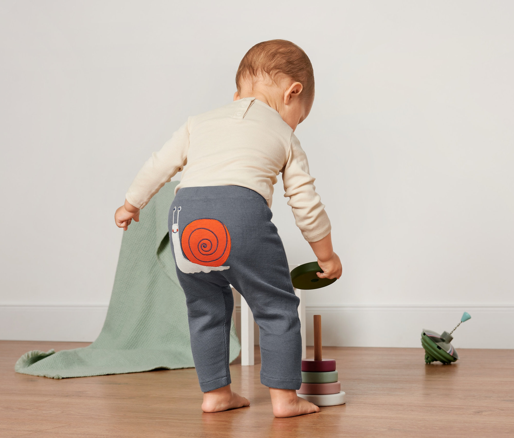 Kleinkind steht mit dem Rücken zur Kamera und trägt eine Baby-Waffelstrukturhose mit Schneckenapplikation.