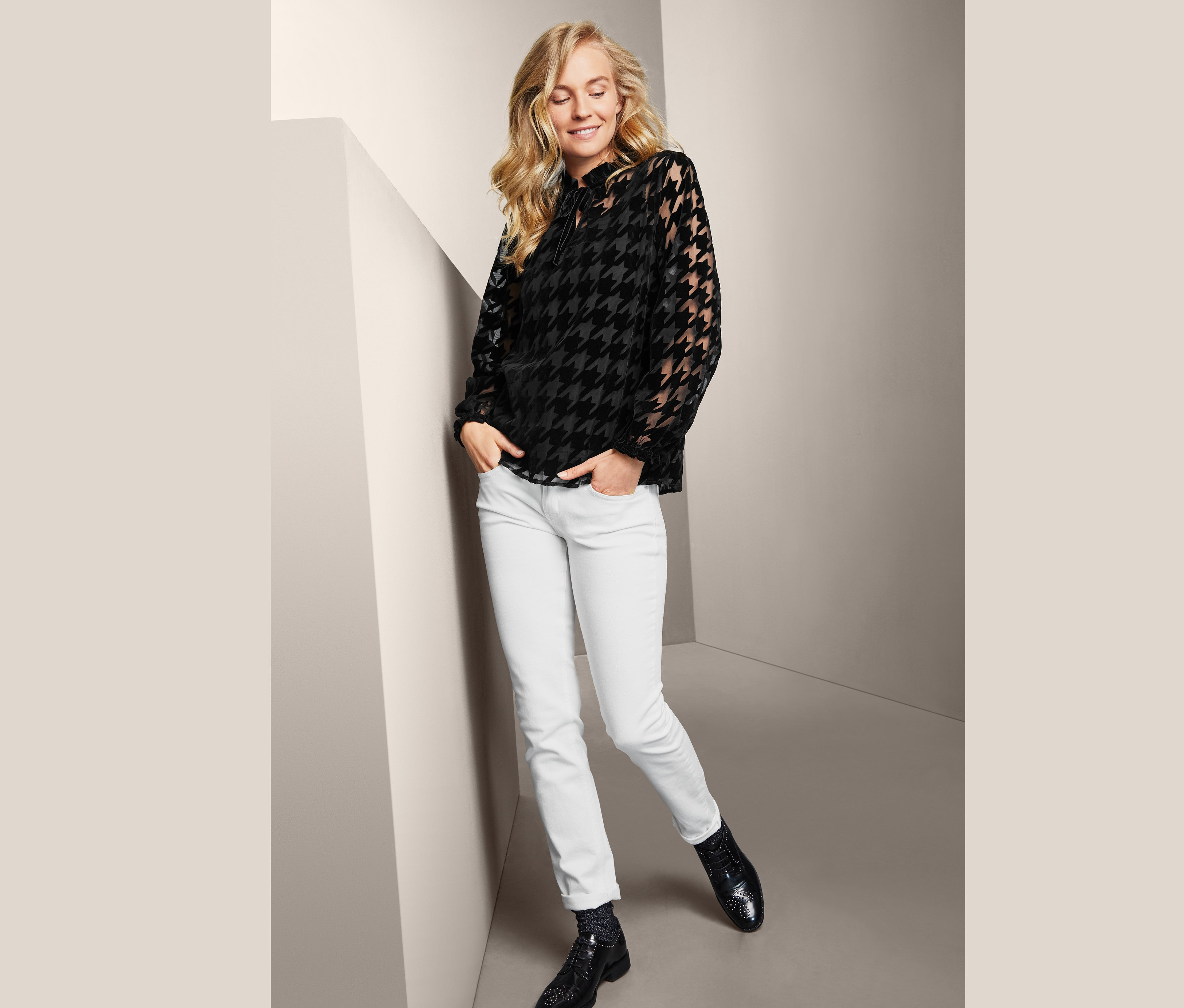 Blonde Frau posiert in Mesh-Bluse mit Hahnentrittmuster, weißen Jeans und schwarzen Schuhen.