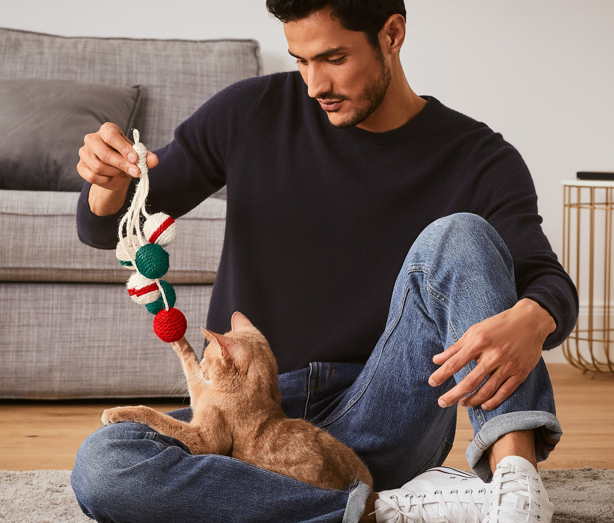 Mann sitzt auf dem Boden und spielt mit einer Katze mit dem Katzenspielzeug »Rassel«.