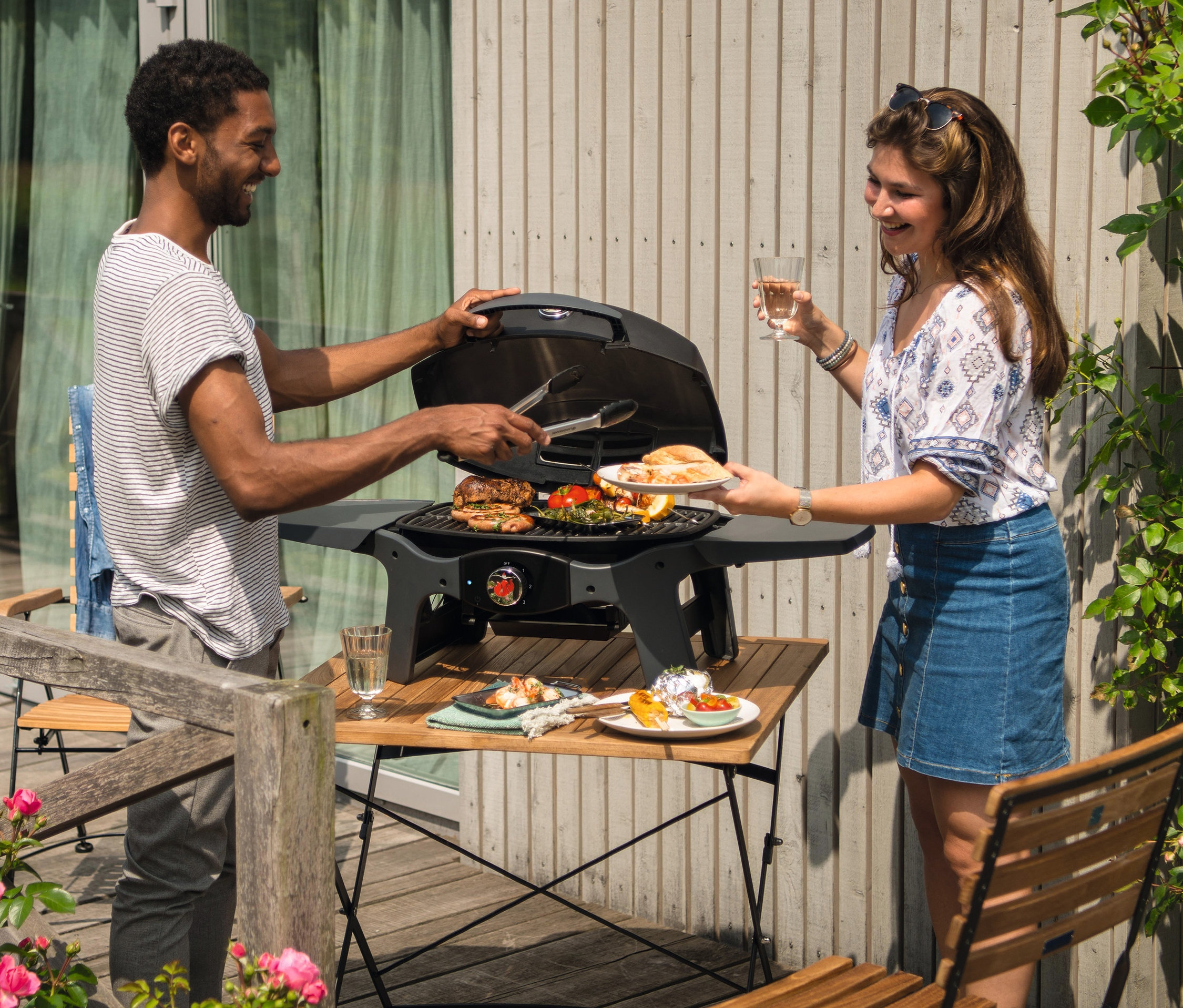 Mann verwendet Grillzange auf Landmann Elektrogrill »12975 Pantera 2.000 W«, während Frau Teller mit Essen und Weinglas hält.