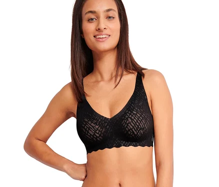 Lächelnde Frau trägt einen schwarzen sloggi ZERO Feel Bliss Soft BH und passende Unterwäsche mit Muster, die Hand an der Hüfte.