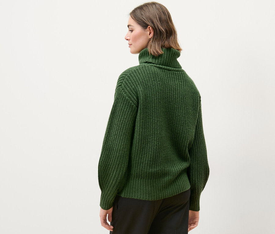 Frau trägt einen grünen Grobstrickpullover mit Rollkragen und schwarze Hosen.