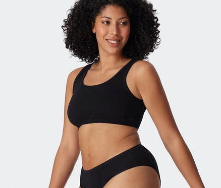 Lächelnde Frau mit dunklem, lockigem Haar trägt ein schwarzes SCHIESSER Bustier und einen schwarzen Slip.