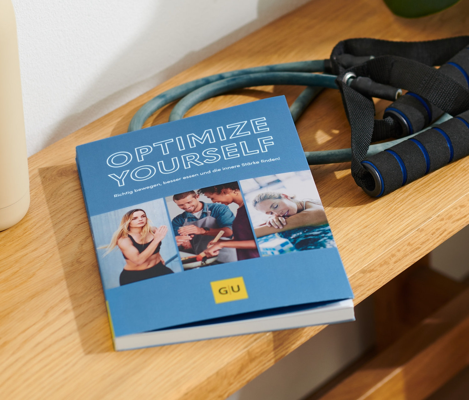 Auf einer Holzbank liegen das Buch »Optimize Yourself«, eine beige Trinkflasche und ein Expander.