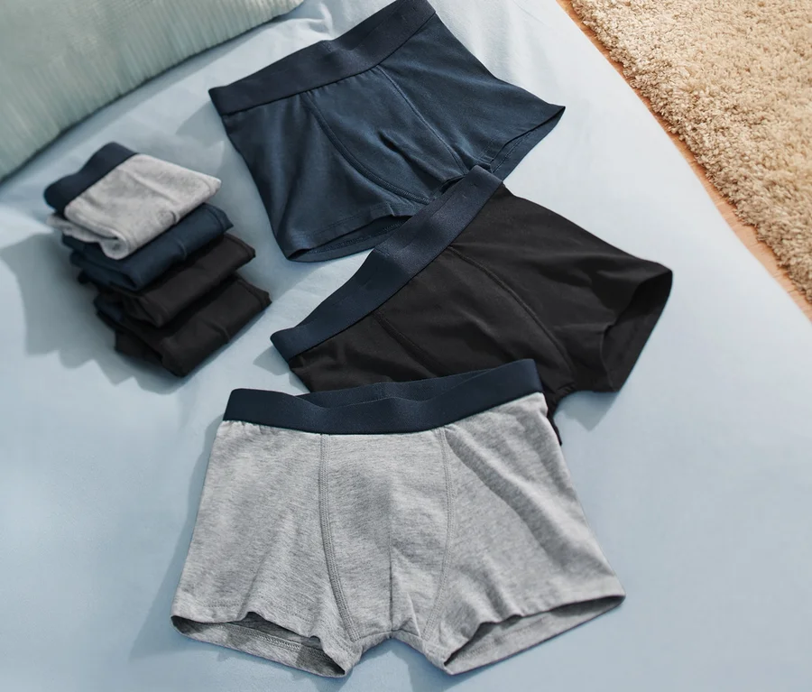 Auf einem hellblauen Bett liegen 7 Kinder-Slipboxer in Grau, Schwarz und Blau, sowohl gestapelt als auch einzeln.