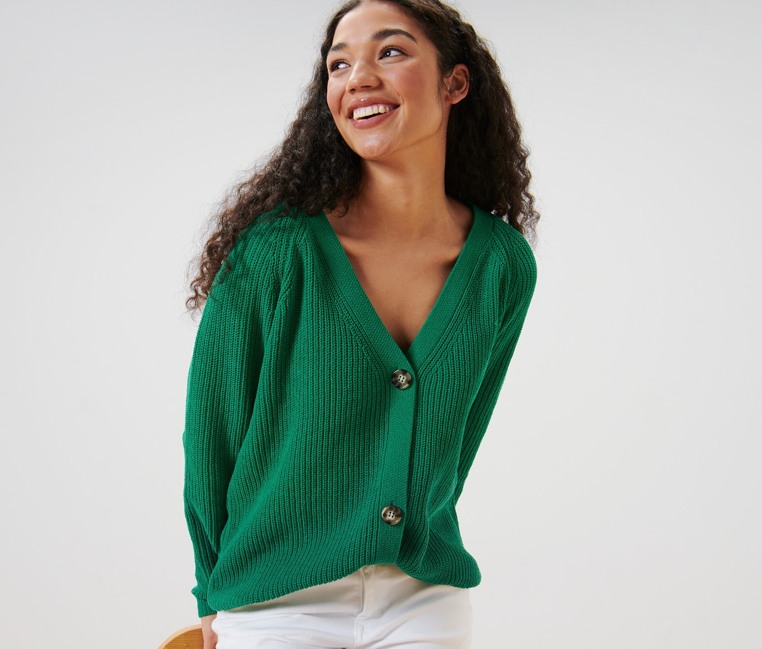 Frau mit lockigem Haar trägt einen grünen Grobstrick-Cardigan und weiße Hose.