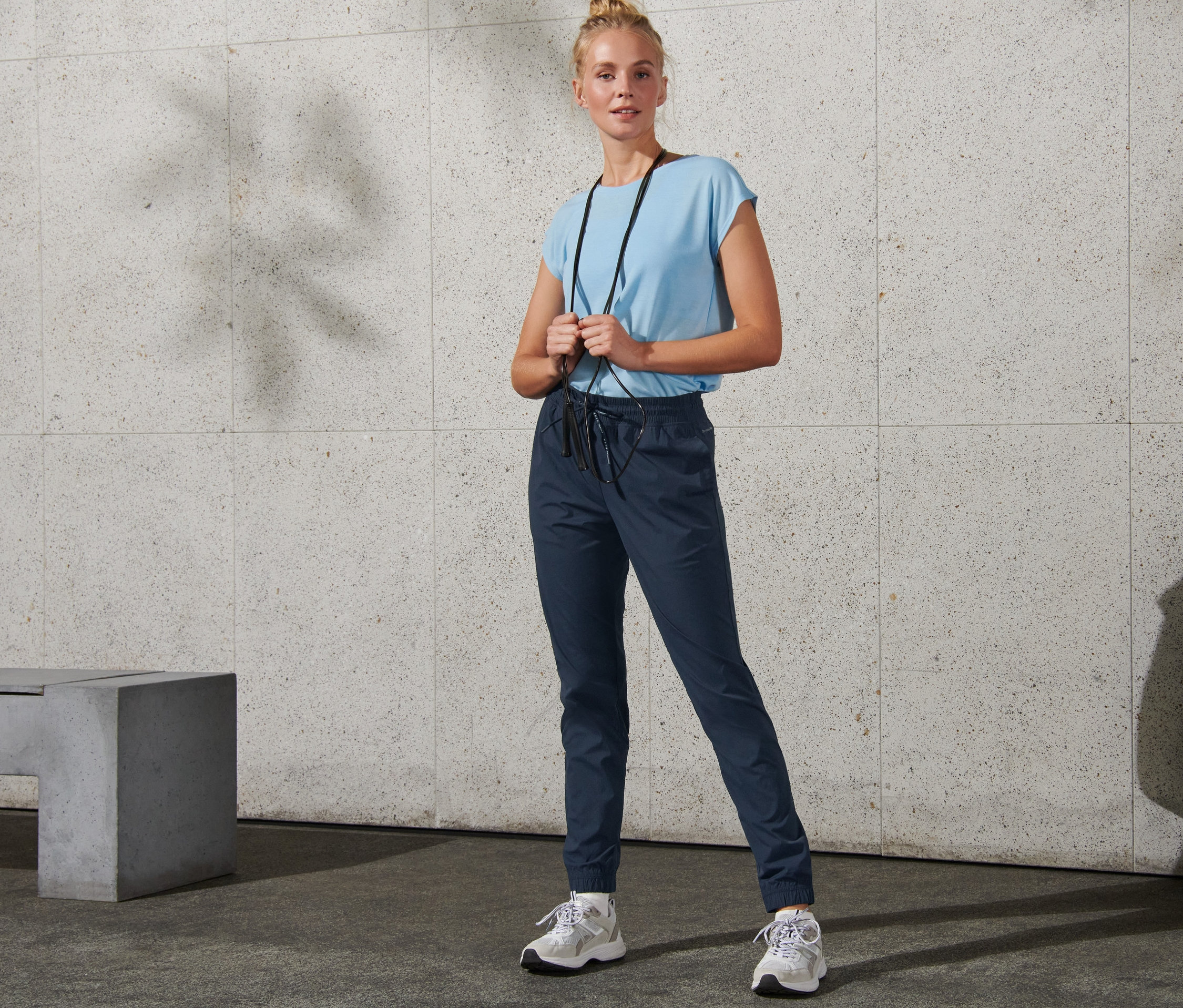 Frau steht mit einem Springseil um den Hals und trägt eine Softshell-Trainingshose.