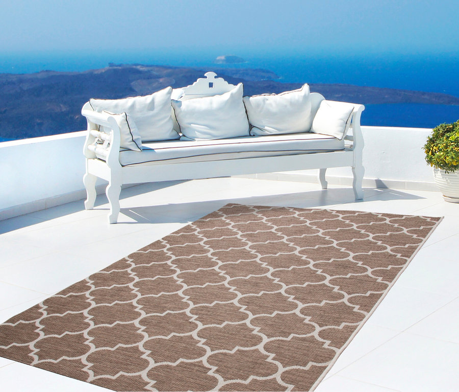 Ein beiger Outdoor-Teppich mit weißem Muster liegt auf einer weißen Terrasse mit einer weißen Bank und Blick auf das Meer.
