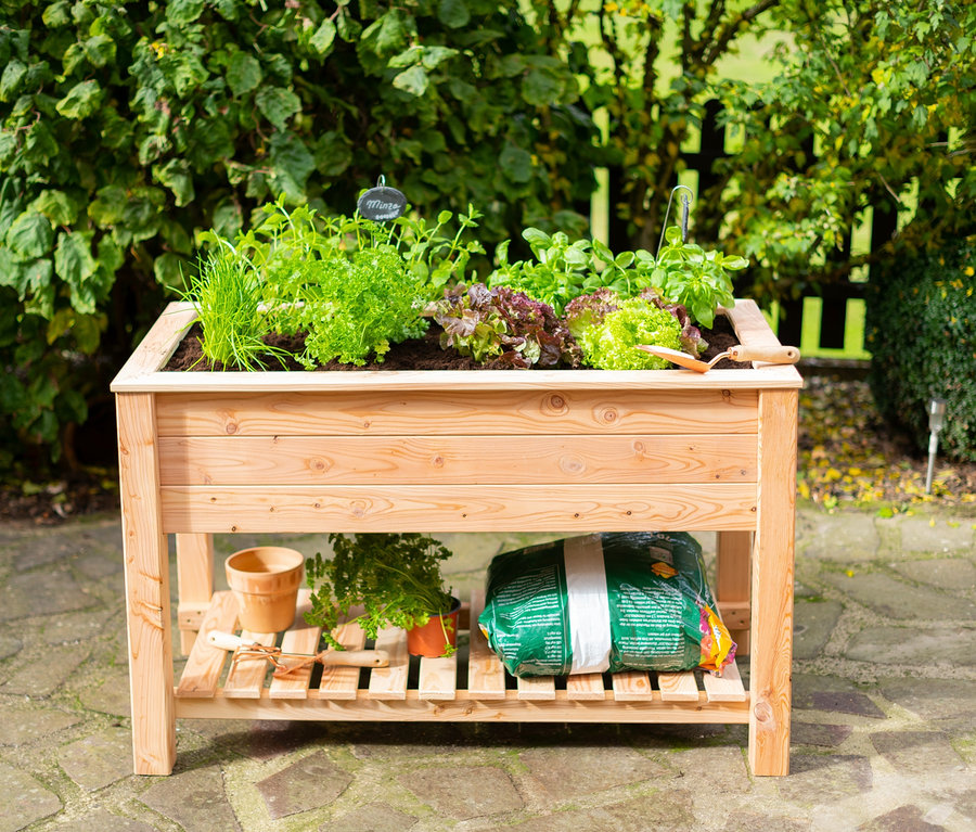 Ein hölzernes Stelzenhochbeet mit verschiedenen Kräutern und Salaten. Darunter befindet sich ein Regal mit einem Blumentopf, einer Pflanze und einem Sack Erde.