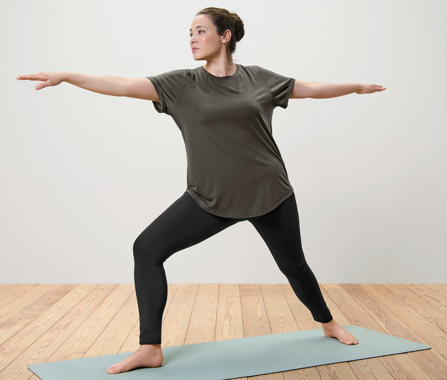 Frau macht Yoga auf einer Matte, sie trägt ein olivgrünes Longshirt und Sporttight.