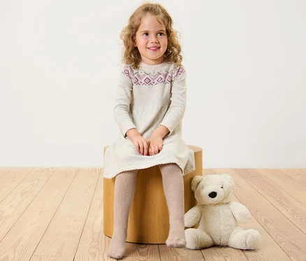 Mädchen sitzt auf einem Holzhocker und trägt ein Kleid und eine Kinder-Strumpfhose mit RWS-Wolle.