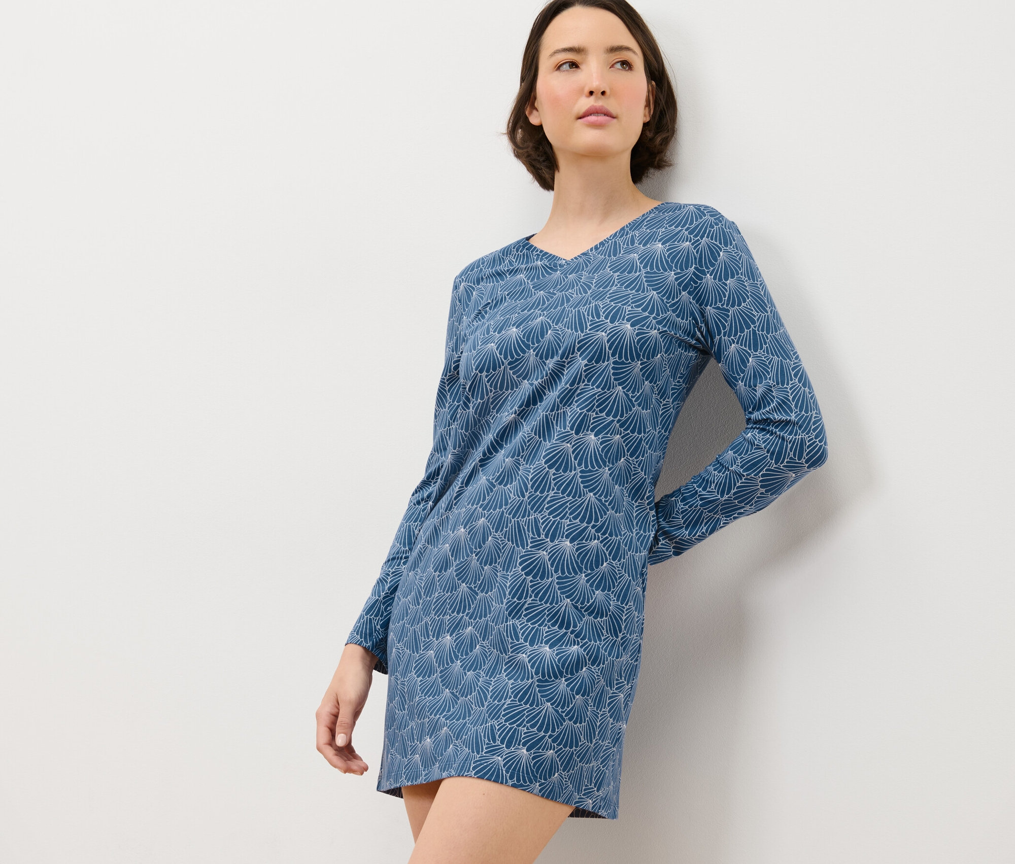 Frau lehnt an einer weißen Wand und trägt ein blau-weiß gemustertes Nachthemd mit langen Ärmeln.