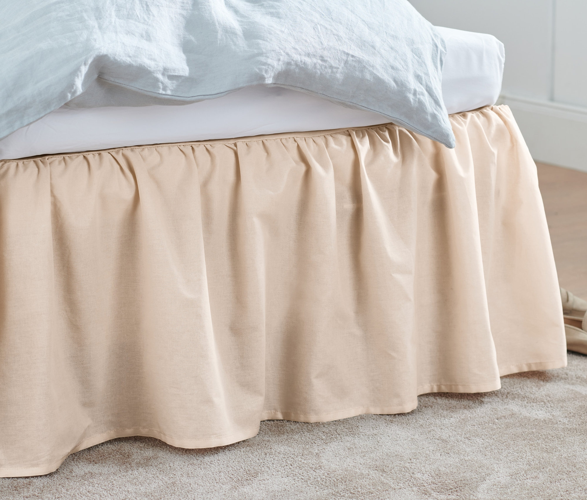 Ein beiger Betthusse verdeckt ein Bett. Beigefarbene Schuhe und ein grauer Teppich liegen auf dem Boden.