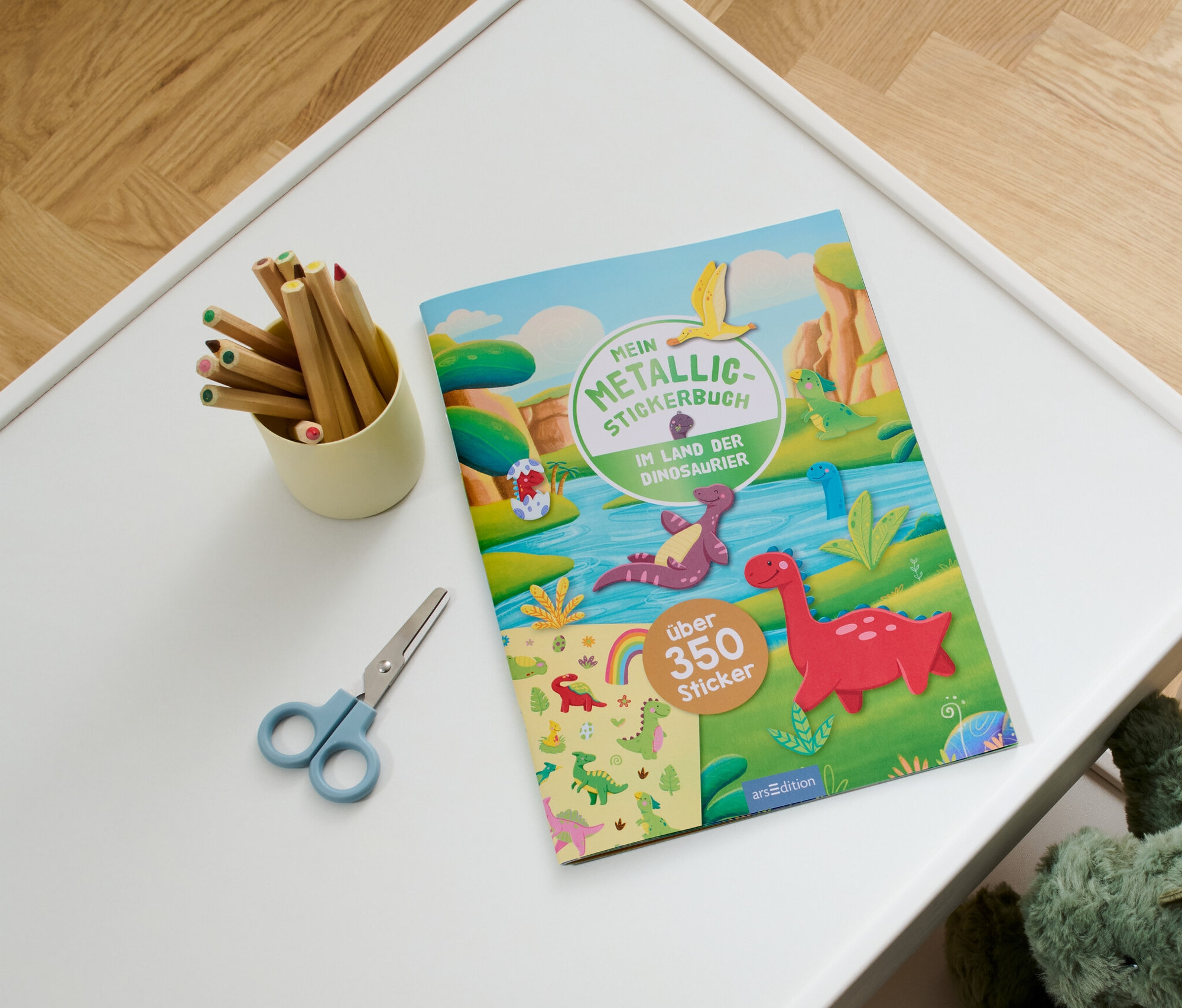 Auf einem weißen Tisch liegen Stifte in einem Becher, eine Schere und das Metallic-Stickerbuch "Im Land der Dinosaurier".