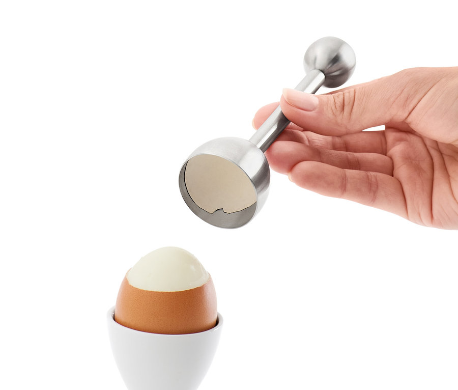 Eine Hand hält einen Eierköpfer über ein Ei in einem weißen Eierbecher.