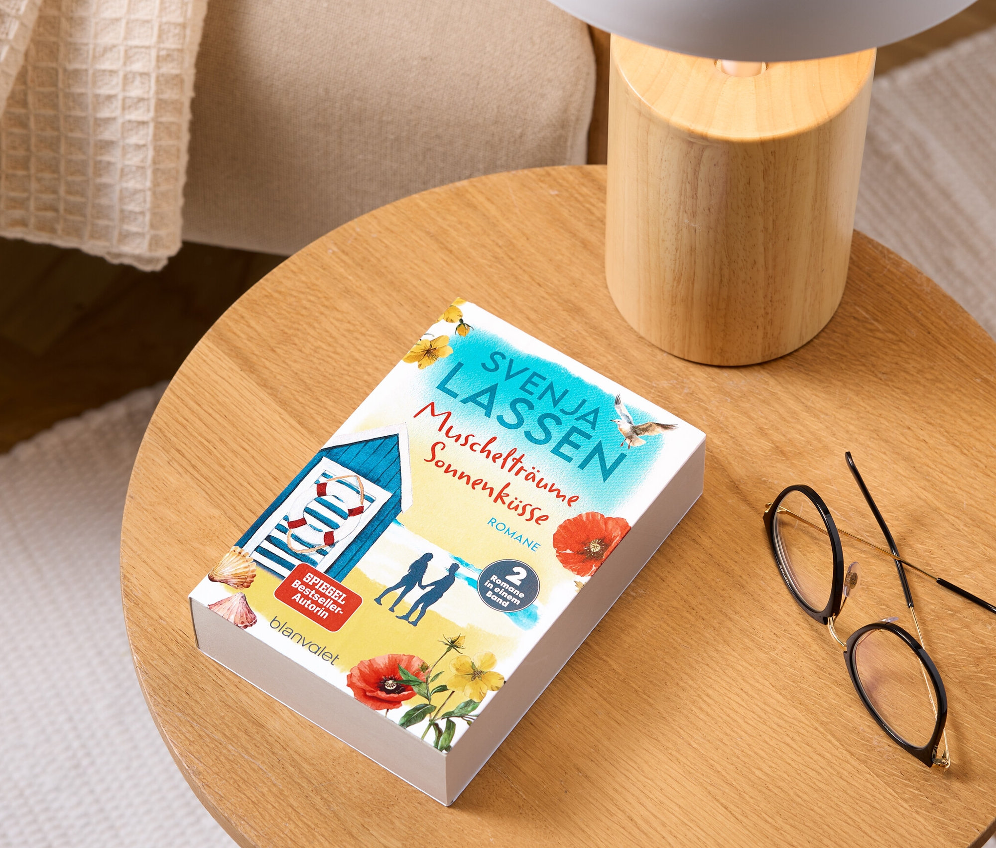 Auf einem Holztisch liegen der Doppelroman »Muschelträume und Sonnenküsse«, eine Lampe und eine Brille.