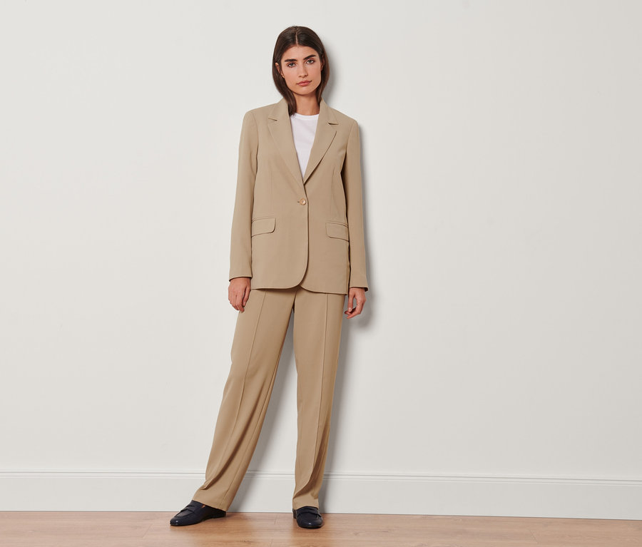 Eine Frau posiert in einem beigen Blazer und einer Webhose mit Biese für die Kamera.