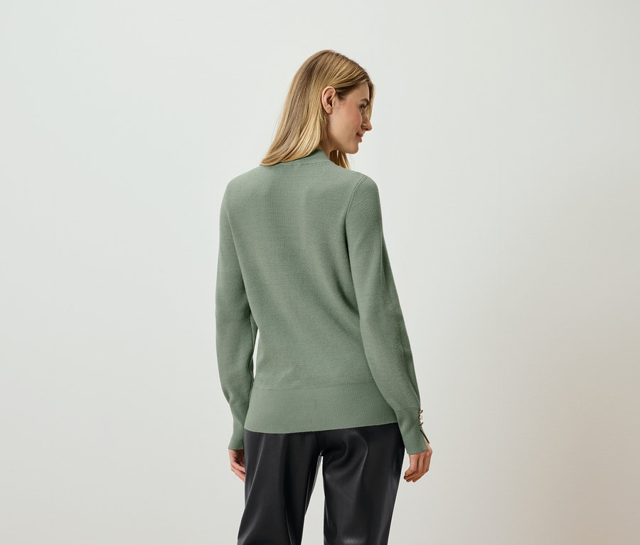 Eine Frau steht mit dem Rücken zur Kamera und trägt einen hellgrünen Pullover und schwarze Hosen.