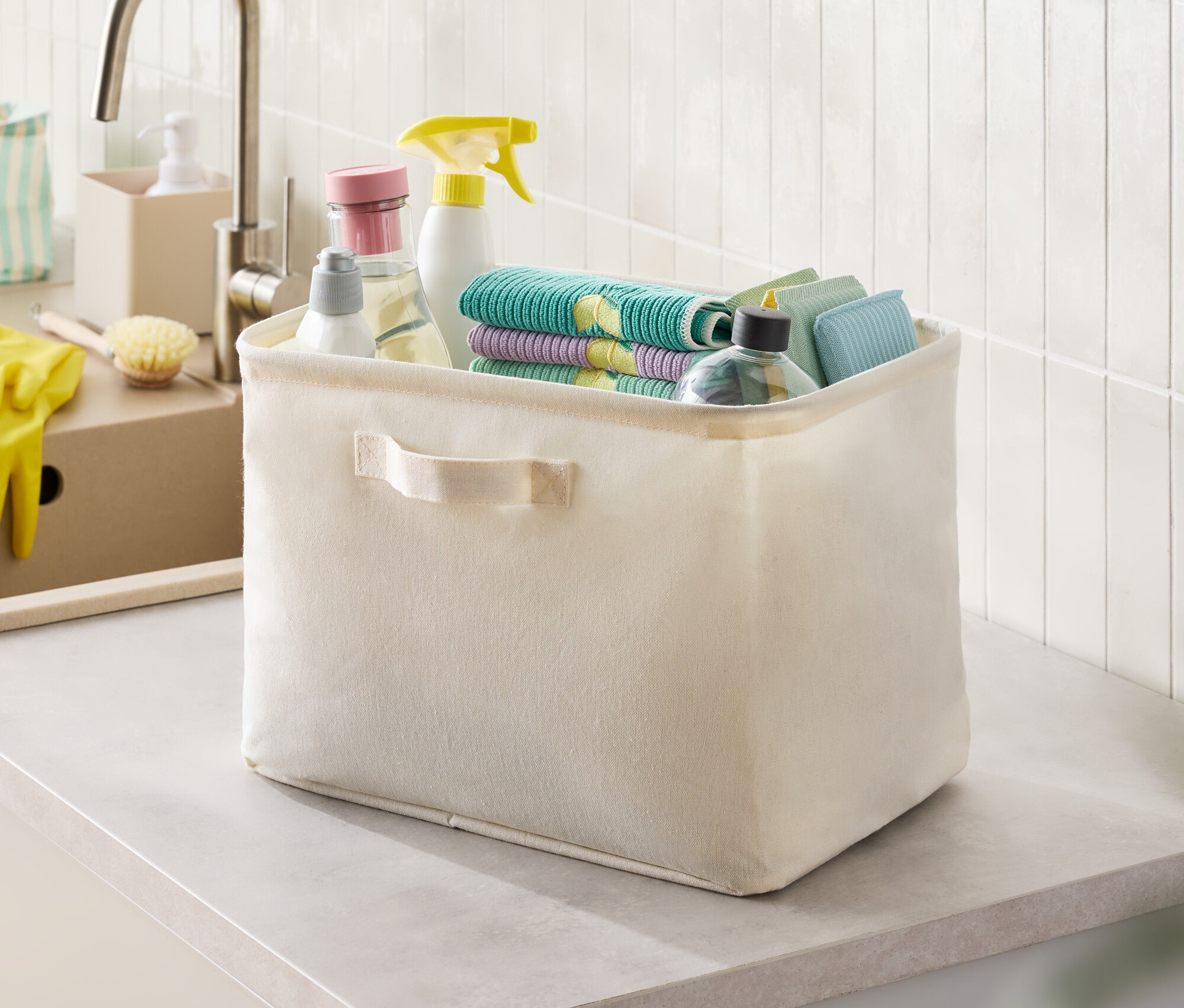Beige Stoffbox mit Putzmitteln und Lappen auf einer Arbeitsplatte.