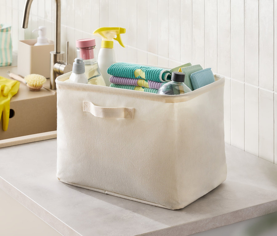 Beige Stoffbox mit Putzmitteln und Lappen auf einer Arbeitsplatte.