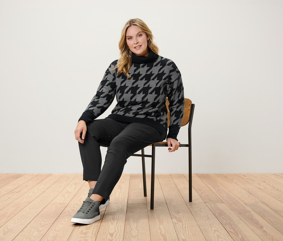 Frau sitzt auf einem Stuhl und trägt einen Hahnentrittpullover, eine schwarze Hose und graue Turnschuhe.