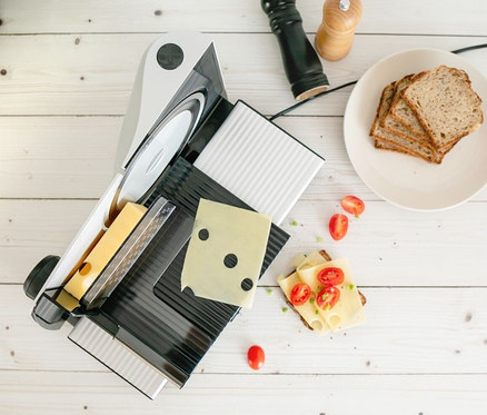 Auf einem weißen Holztisch stehen ein Steba Allesschneider »AS 200« mit einem Stück Käse, ein Teller mit Brot, Pfeffer und Tomaten.