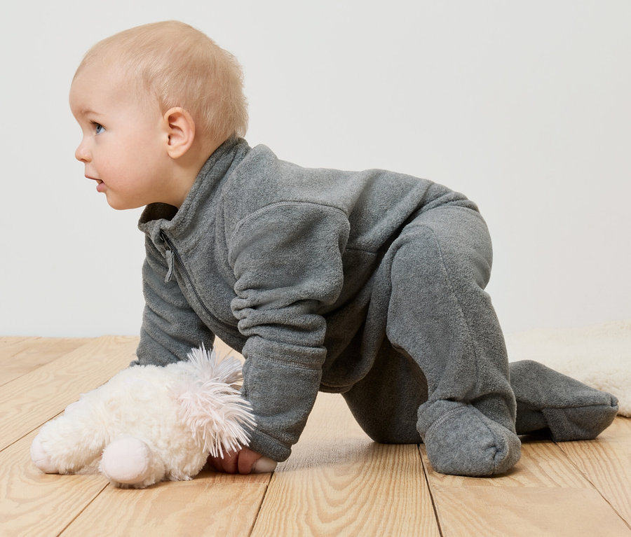 Ein Baby im Fleece-Einteiler krabbelt auf dem Bauch neben einem Stofftier.