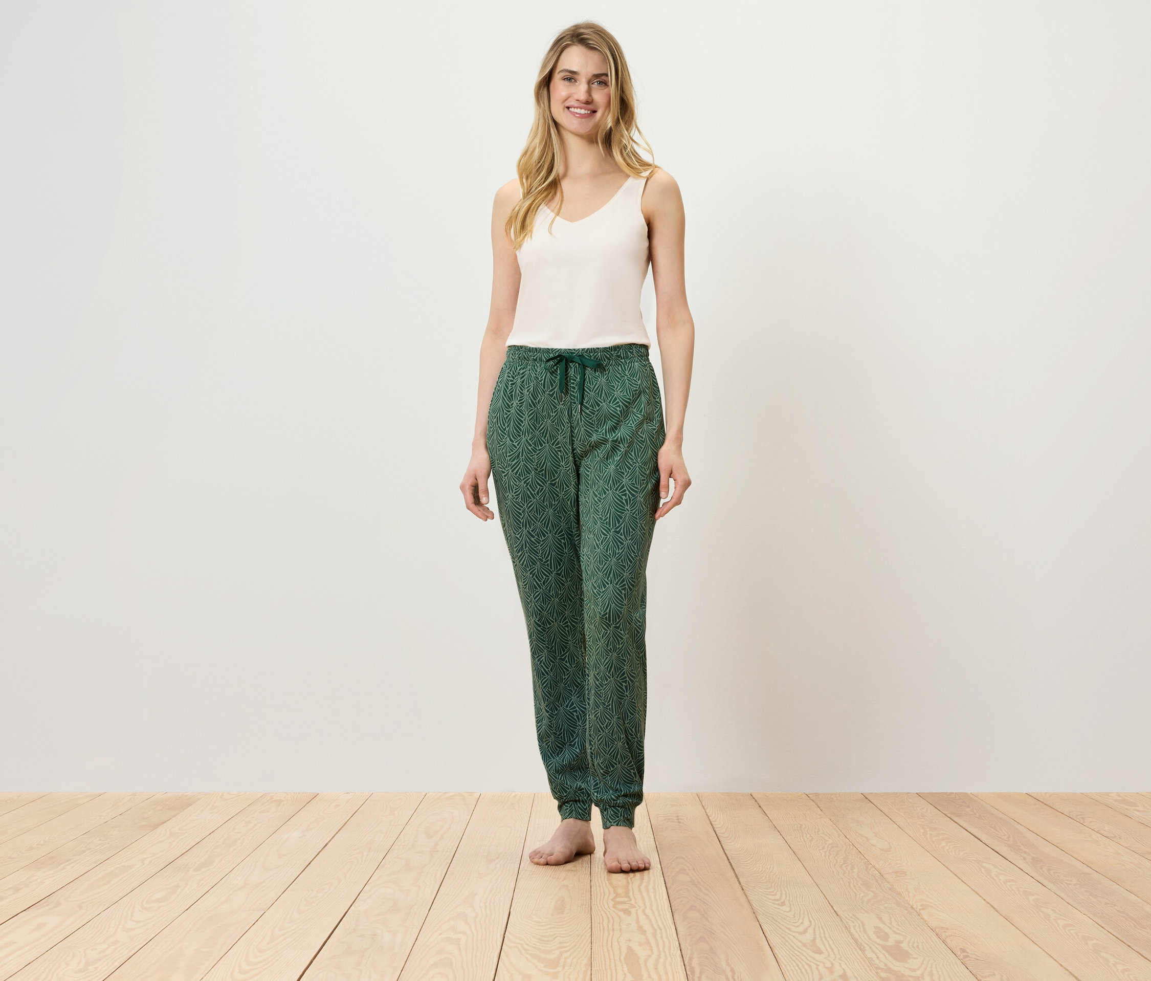 Eine Frau steht auf einem Holzboden und trägt ein weißes Top und eine grüne Hose.