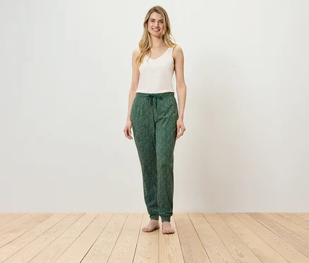 Eine Frau steht auf einem Holzboden und trägt ein weißes Top und eine grüne Hose.