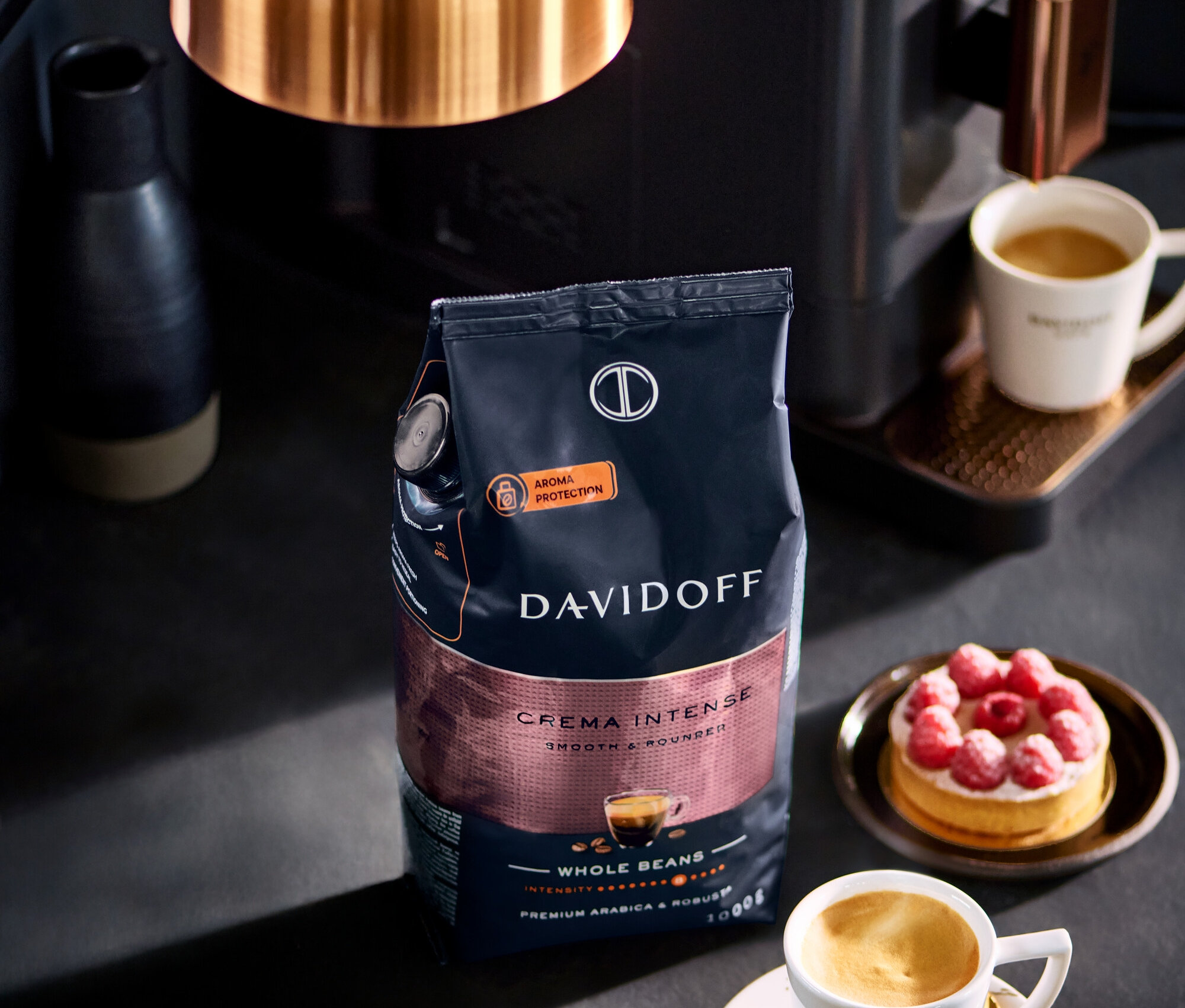 Eine Packung DAVIDOFF CAFÉ Crema Intense 1000 g ganze Bohne steht neben einer Tasse Kaffee und einem Himbeerkuchen.
