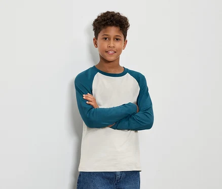 Junge mit braunem lockigem Haar steht mit verschränkten Armen und trägt ein blaugrünes Langarmshirt und Jeans.