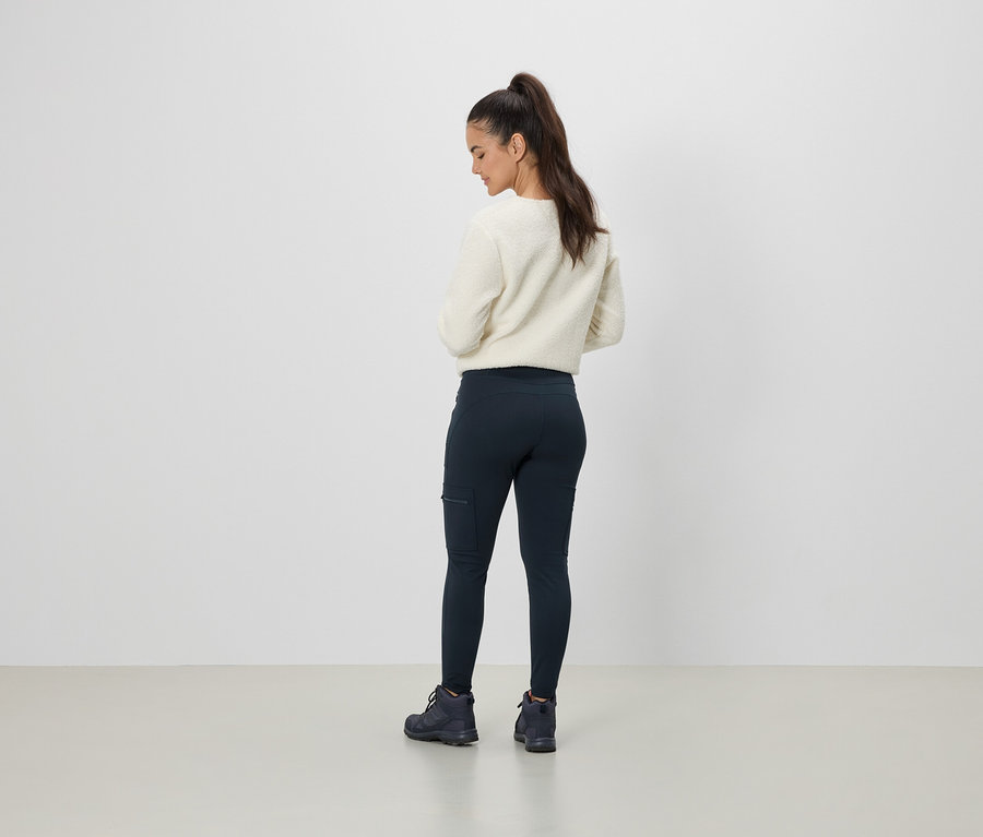 Frau trägt einen cremefarbenen Pullover und dunkelblaue Leggings mit einer Tasche, von hinten gesehen.