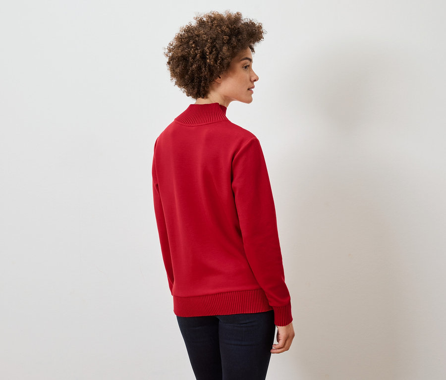 Frau mit lockigem Haar steht seitlich und trägt einen roten Pullover und dunkelblaue Jeans.