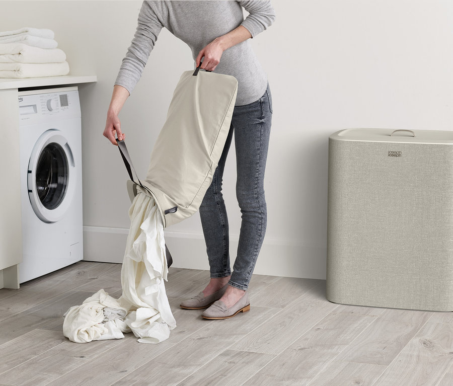 Eine Frau leert weiße Wäsche aus einem beigen Wäschesack auf einen Haufen auf dem Boden. Im Hintergrund sind eine Waschmaschine und ein Wäschekorb zu sehen.